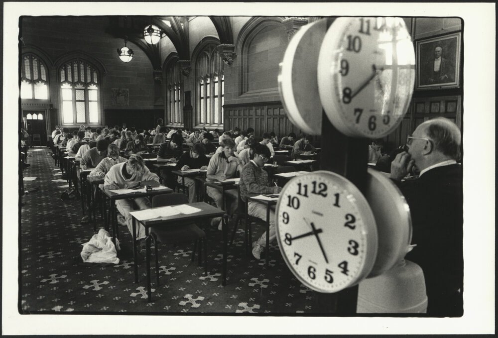 Exam Supervisor Overseeing Students in MacLaurin Hall with Large Clocks Displaying Time to His Left
