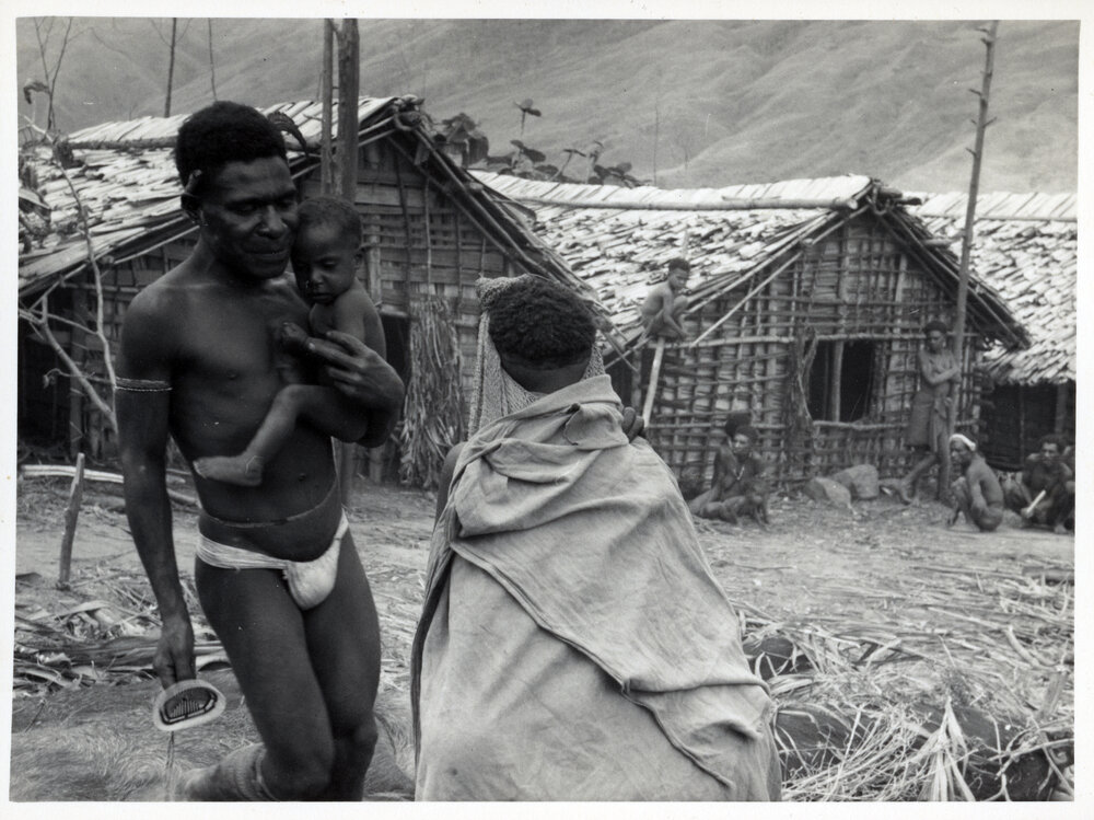 Man Carrying Baby, With Others Seated by Building