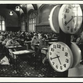 Exam Supervisor Overseeing Students in MacLaurin Hall with Large Clocks Displaying Time to His Left