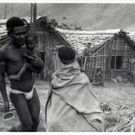 Man Carrying Baby, With Others Seated by Building